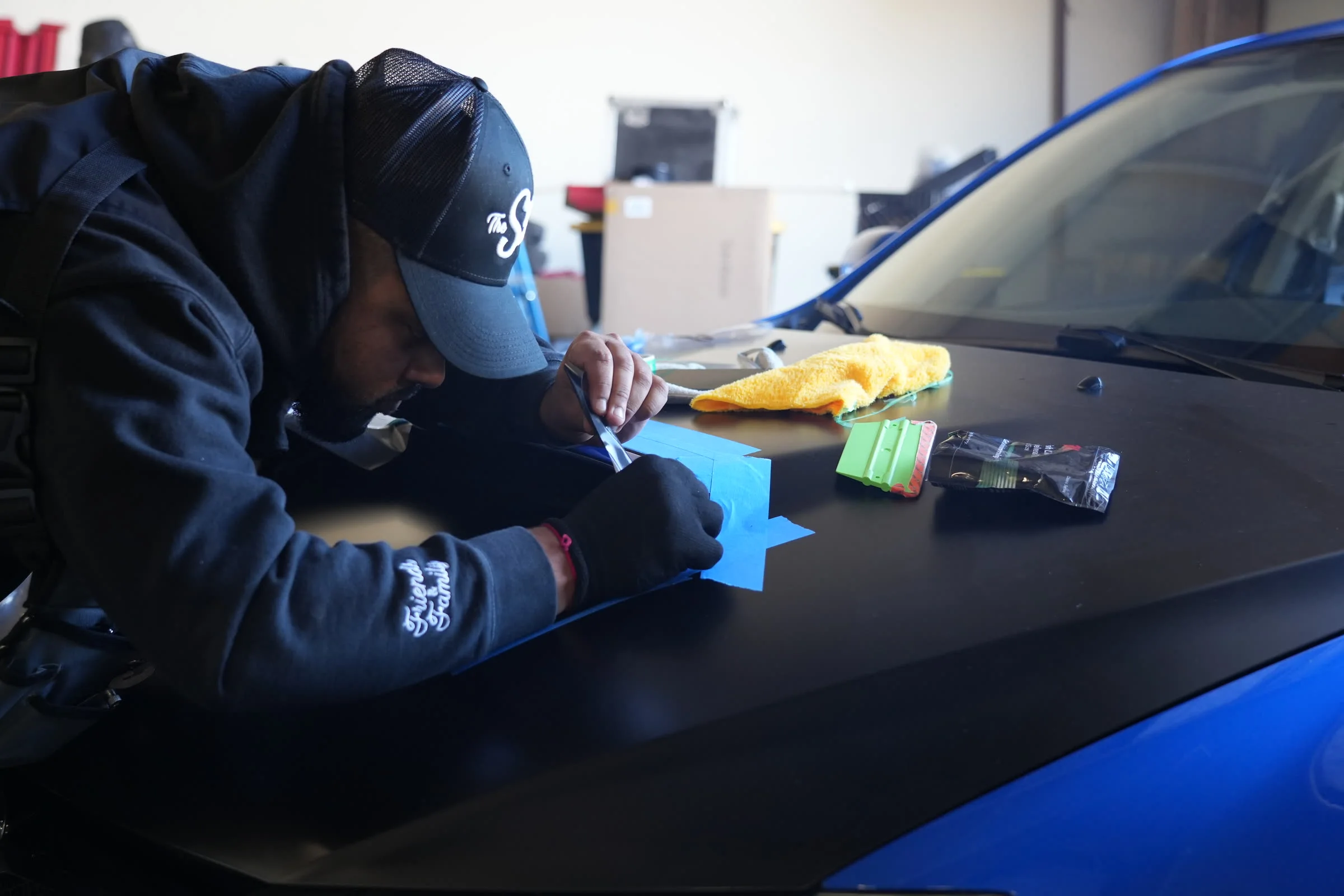 A Sticker Smith installer in a branded cap cutting blue vinyl on a matte car hood